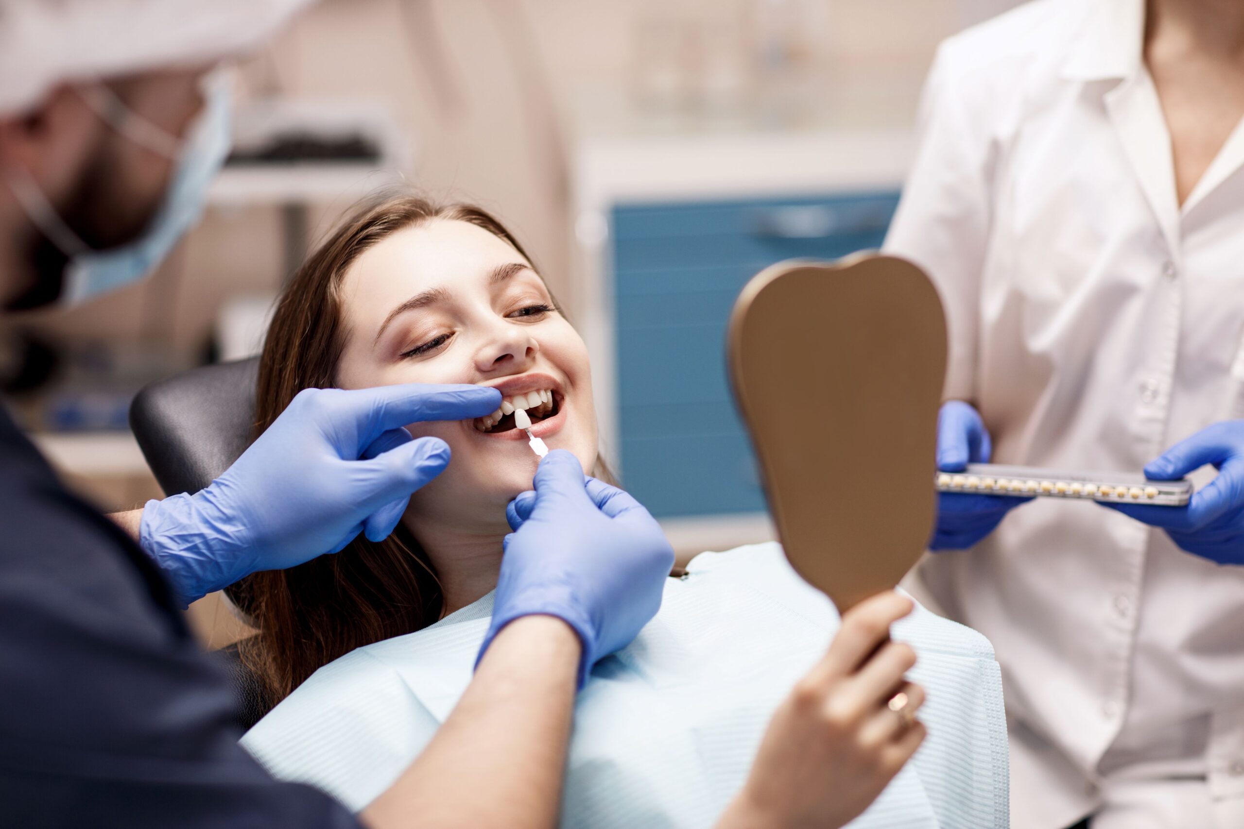 Young woman holding mirror while dentist color-matches her teeth.